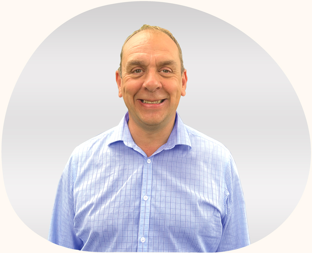 A man with short hair, wearing a pale blue checked shirt, smiles. Behind him is a plain pale grey background