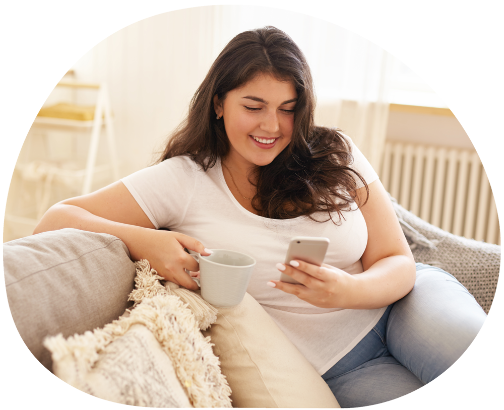 Woman with long dark hair reclines on a cream sofa, smiling while looking at her phone and holding a cream mug.