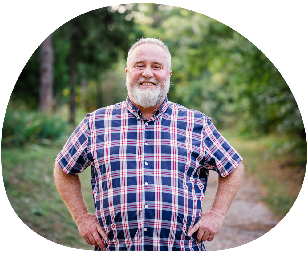 Man with short white hair and a white beard, wearing a checked shirt, standing outside with his hands on his hips while smiling.